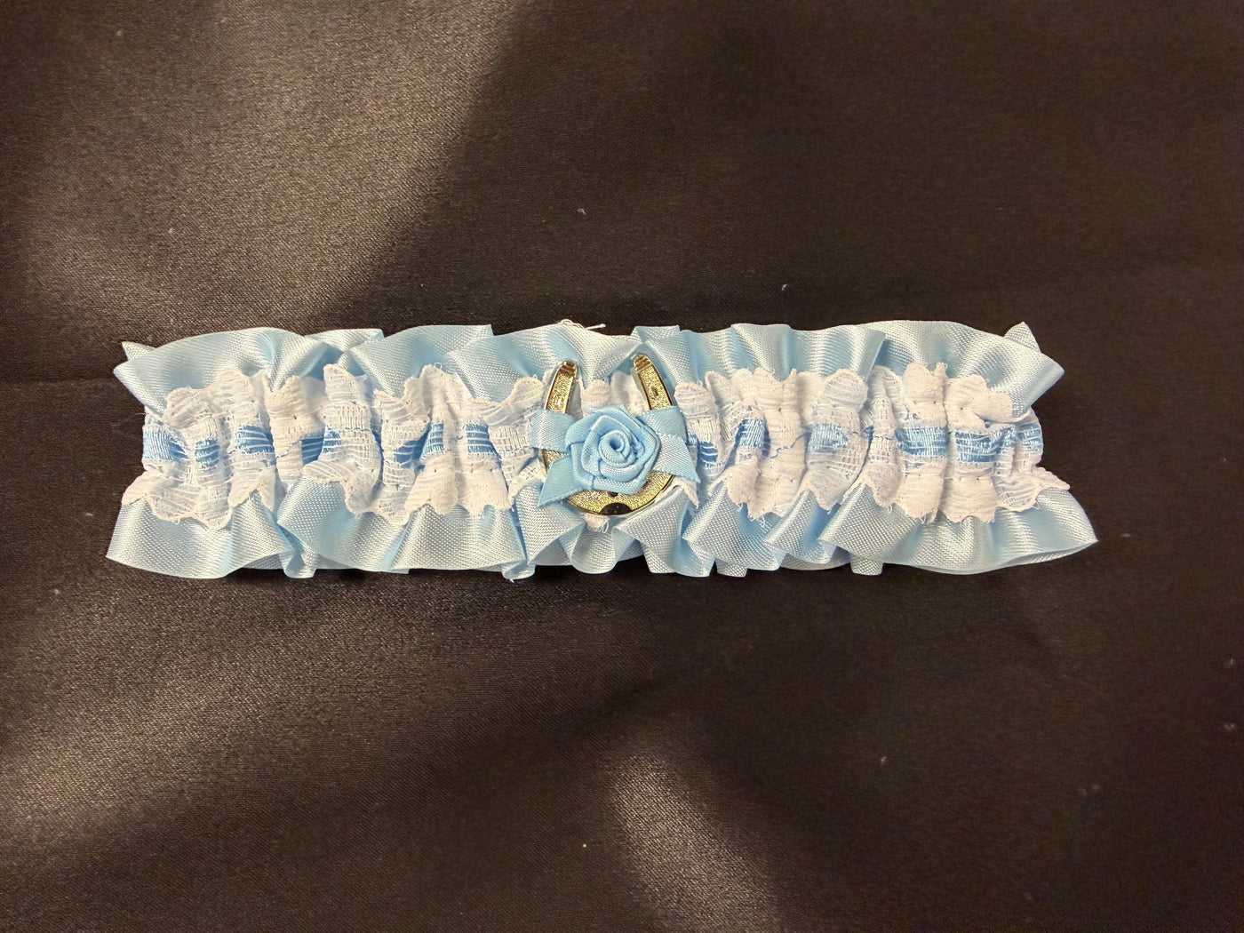 A light blue and white ruffled lace garter belt with a central blue fabric rose and gold-toned metal accents, displayed on a dark brown textured background.